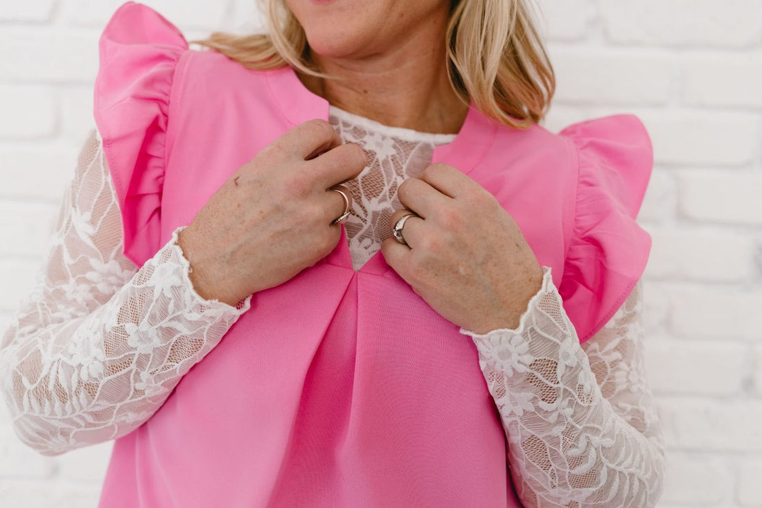 Person in a pink top with lace sleeves adjusting their outfit against a white brick wall.
