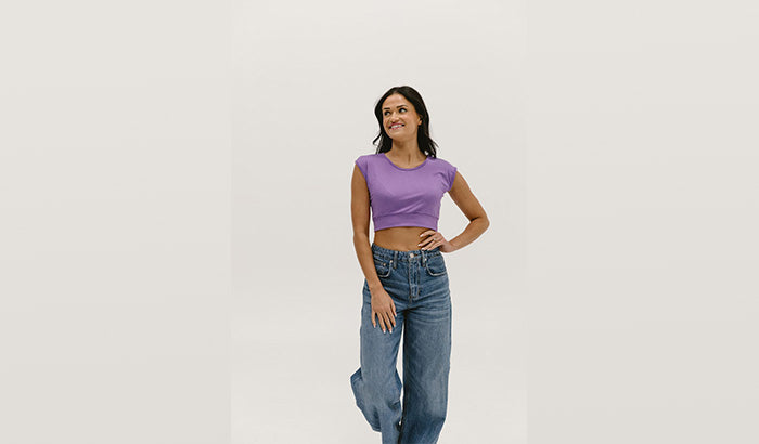 Woman in a purple crop top and jeans posing against a white background.