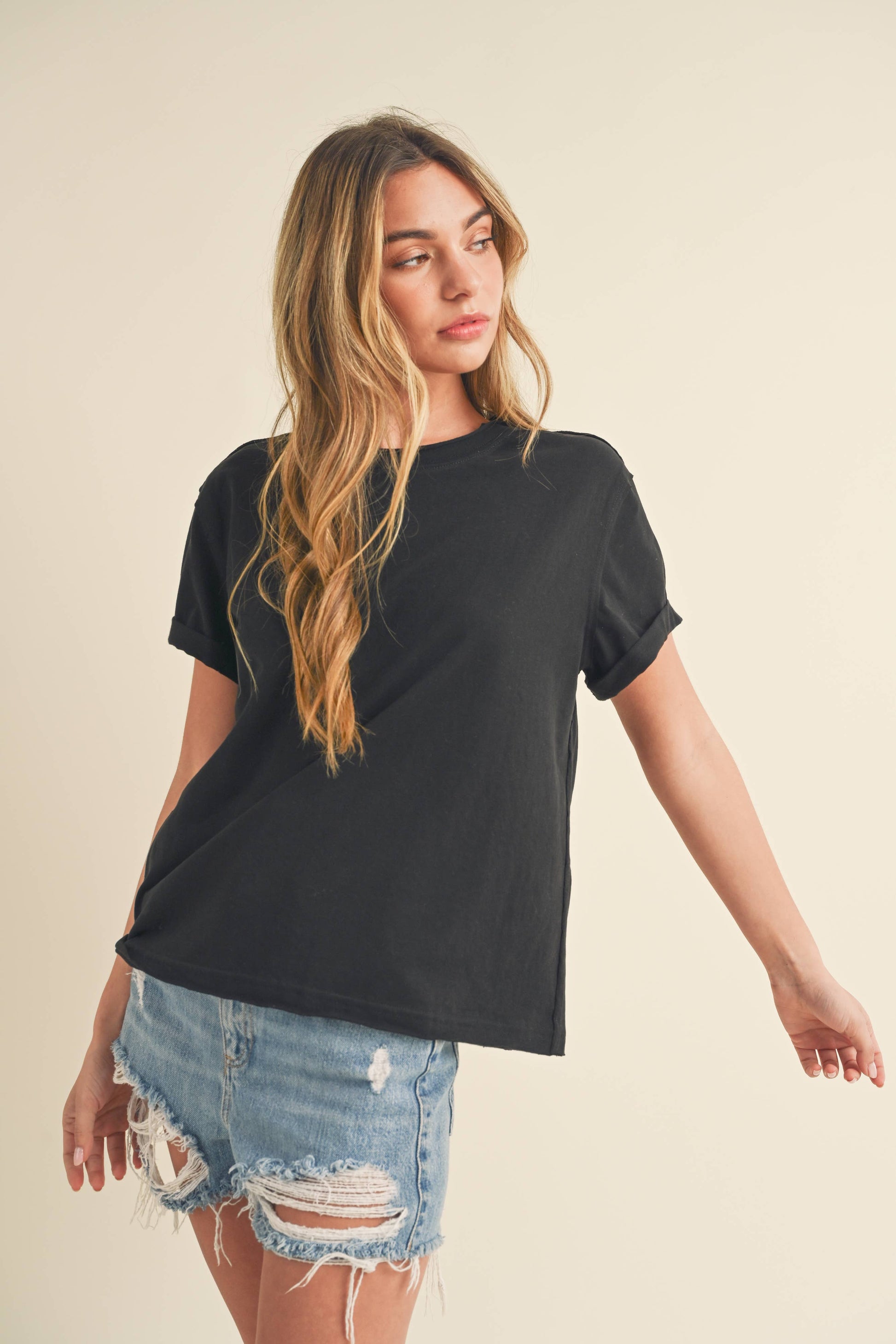 Young woman in a black T-shirt and distressed denim shorts stands against a plain background, looking off to the side with a relaxed pose.