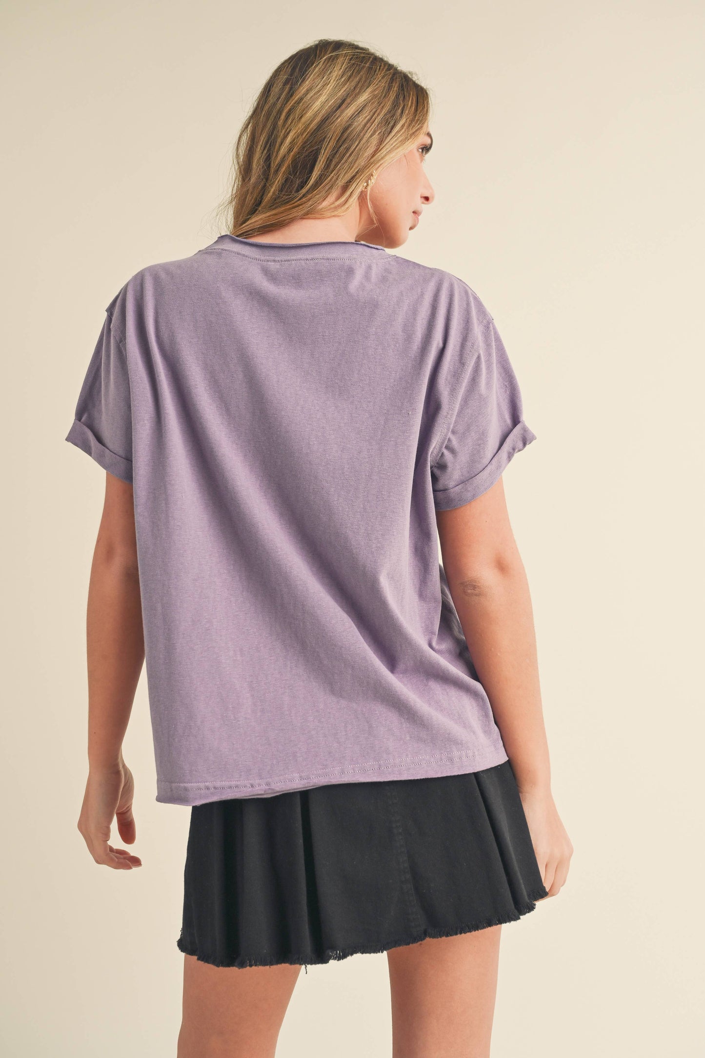 A woman with long hair stands facing away, wearing a lavender t-shirt and a black skirt. The neutral background highlights a casual, relaxed mood.