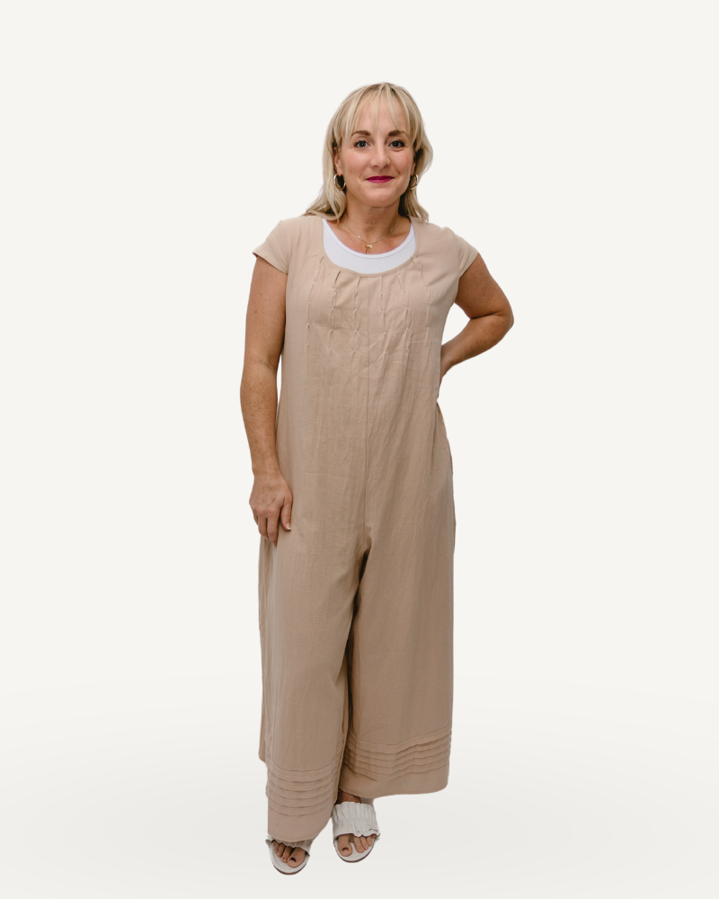 A woman in a beige jumpsuit, with short sleeves, stands confidently.