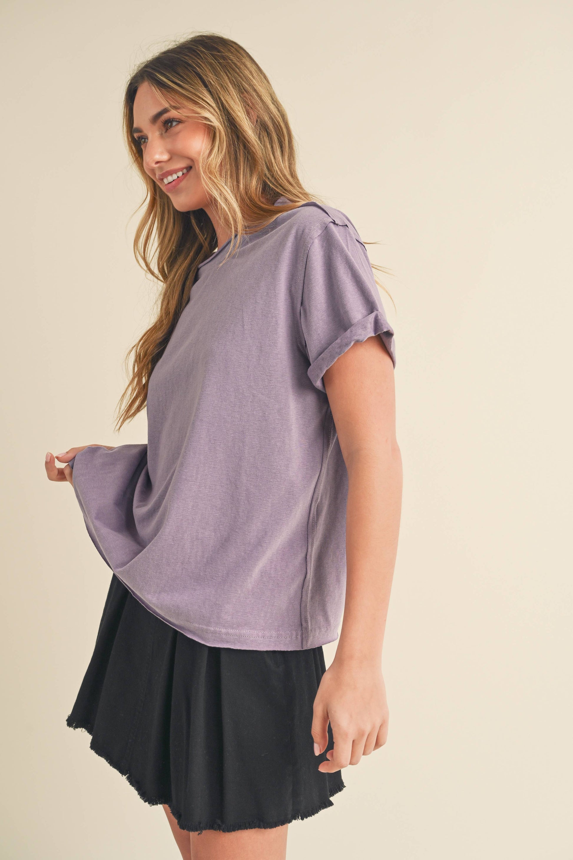 Smiling woman in a purple T-shirt and black skirt stands sideways against a beige background. Her relaxed posture conveys a casual, happy mood.
