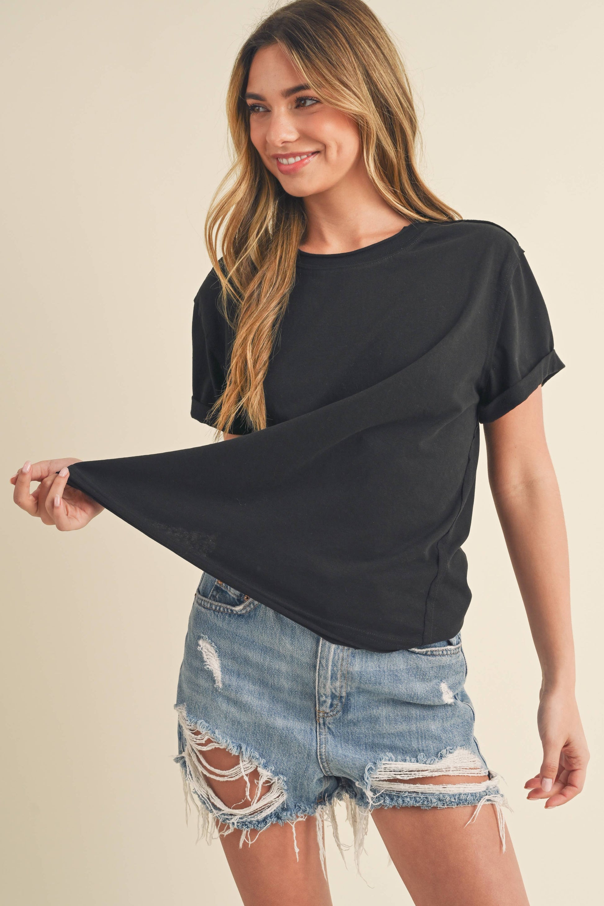 Smiling woman in a black T-shirt and distressed denim shorts holds out her shirt, conveying a casual and relaxed vibe against a neutral background.