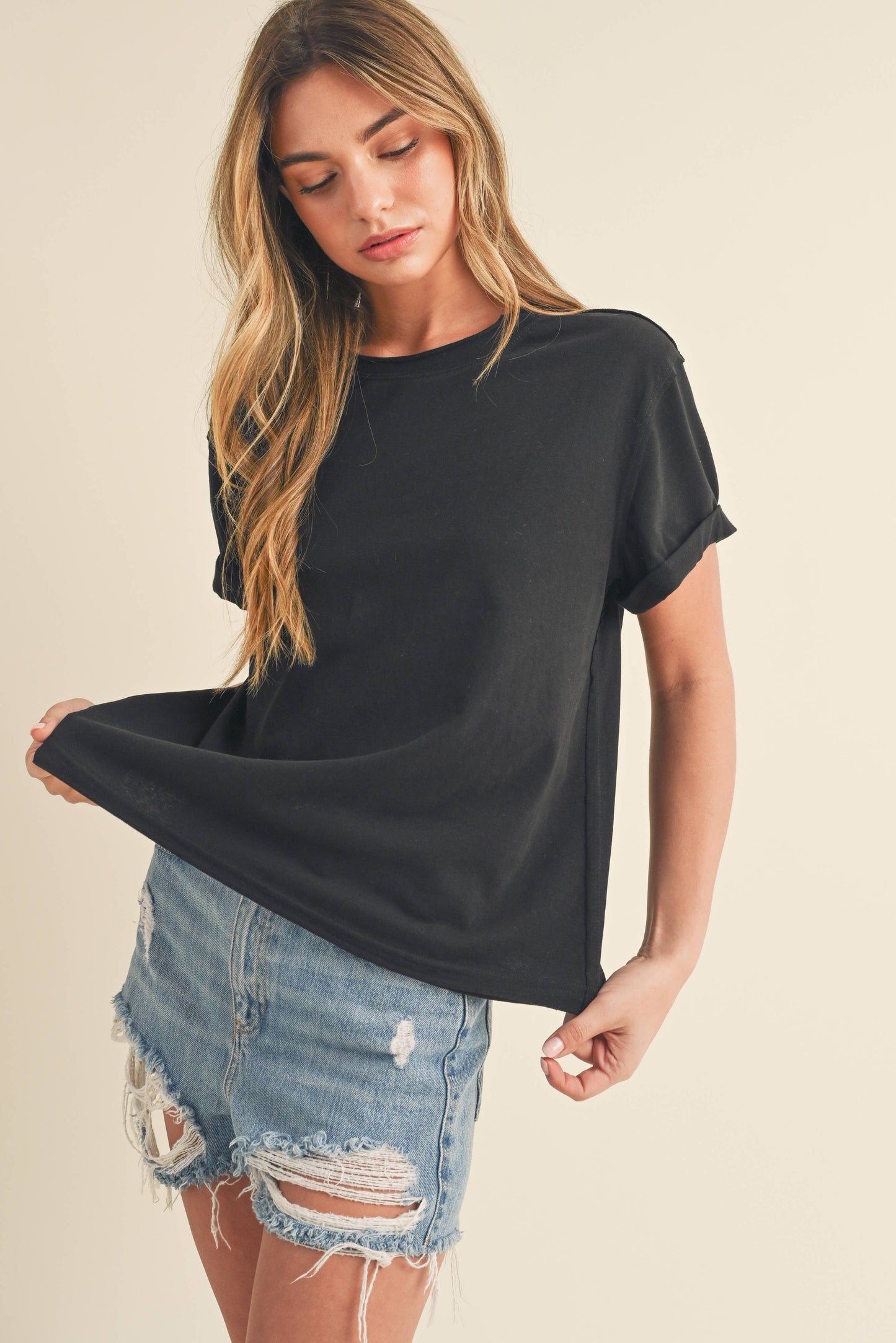 Woman in a black t-shirt and distressed denim shorts looks down, holding the side of her shirt. The setting is neutral, conveying a casual, relaxed vibe.