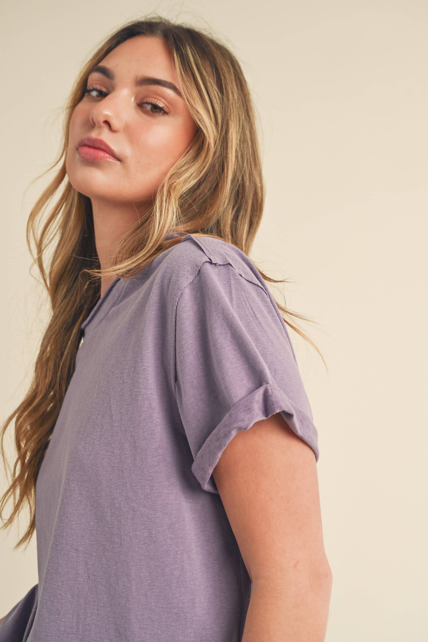 Young woman with long, wavy hair, wearing a lavender t-shirt, looks confidently at the camera. Her expression is calm, and the background is neutral.