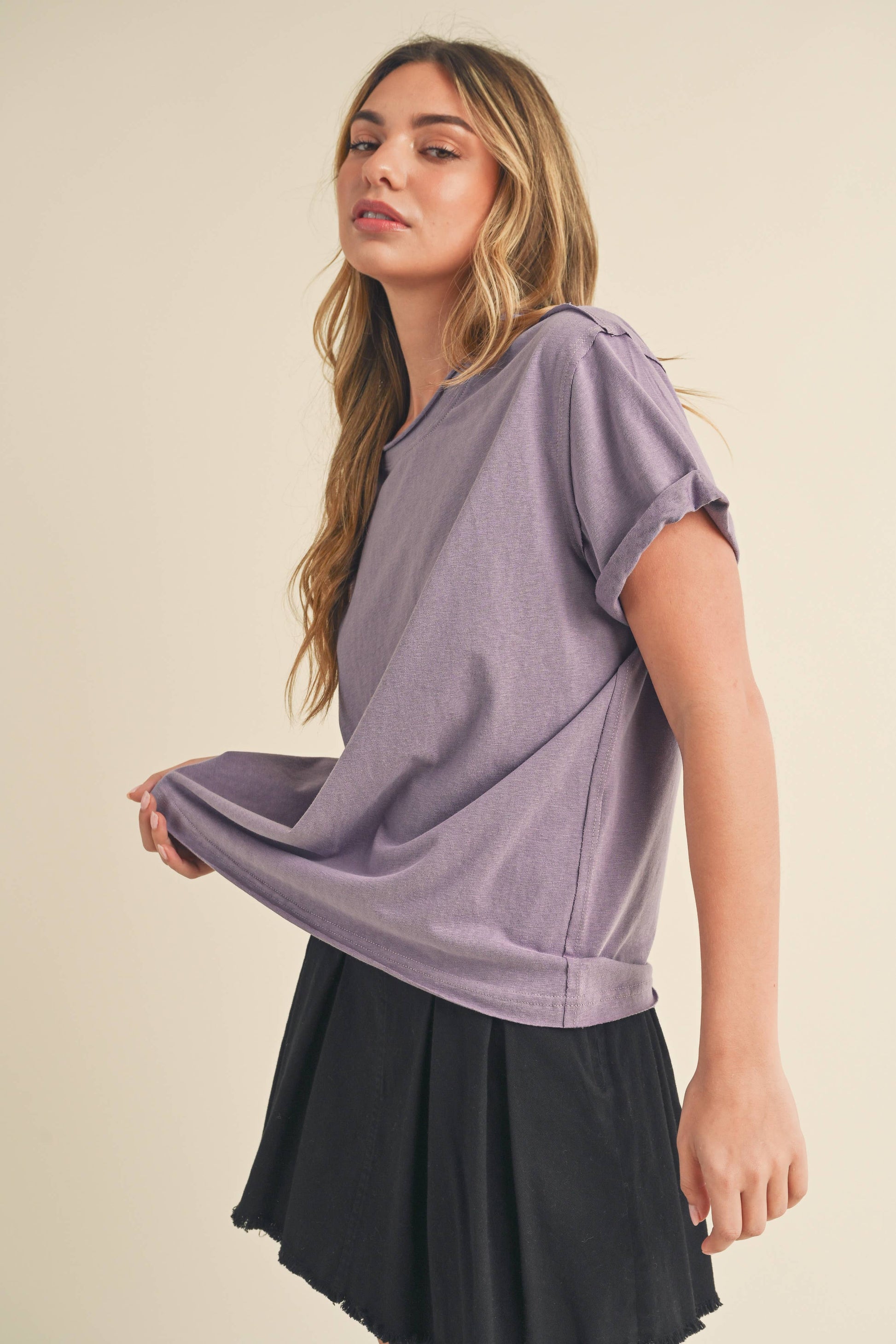 Young woman with long hair wears a casual purple t-shirt and black skirt, slightly pulling the shirt to the side, conveying a relaxed and confident tone.