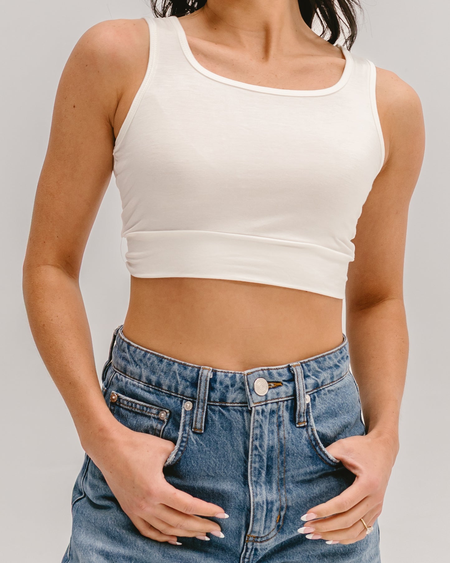A cropped view of a person wearing a white sleeveless Halftee paired with high-waisted light-wash jeans, posing with hands resting in pockets.