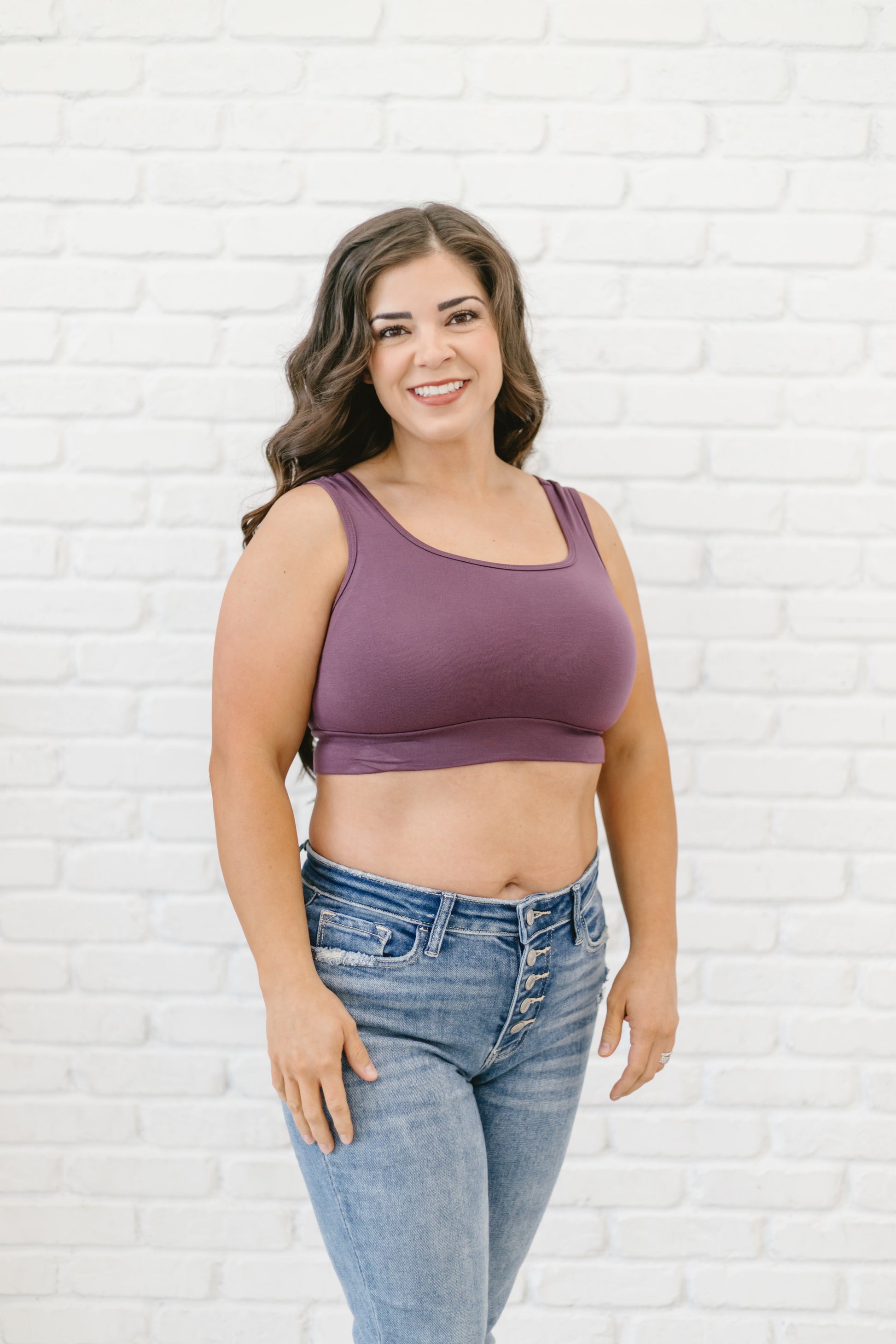 Woman with long dark hair wearing a plum Halftee crop top paired with blue denim jeans, standing in front of a white brick wall.