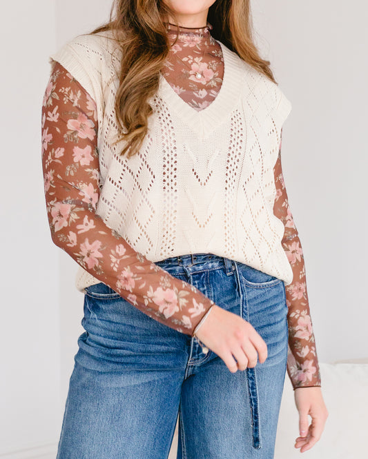 Woman wearing a cream-colored open-knit sweater vest over a brown floral print turtleneck and blue jeans.