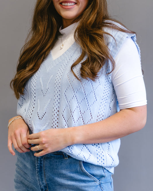 Smiling woman wearing a light blue open-knit sweater vest over a white turtleneck and blue jeans.