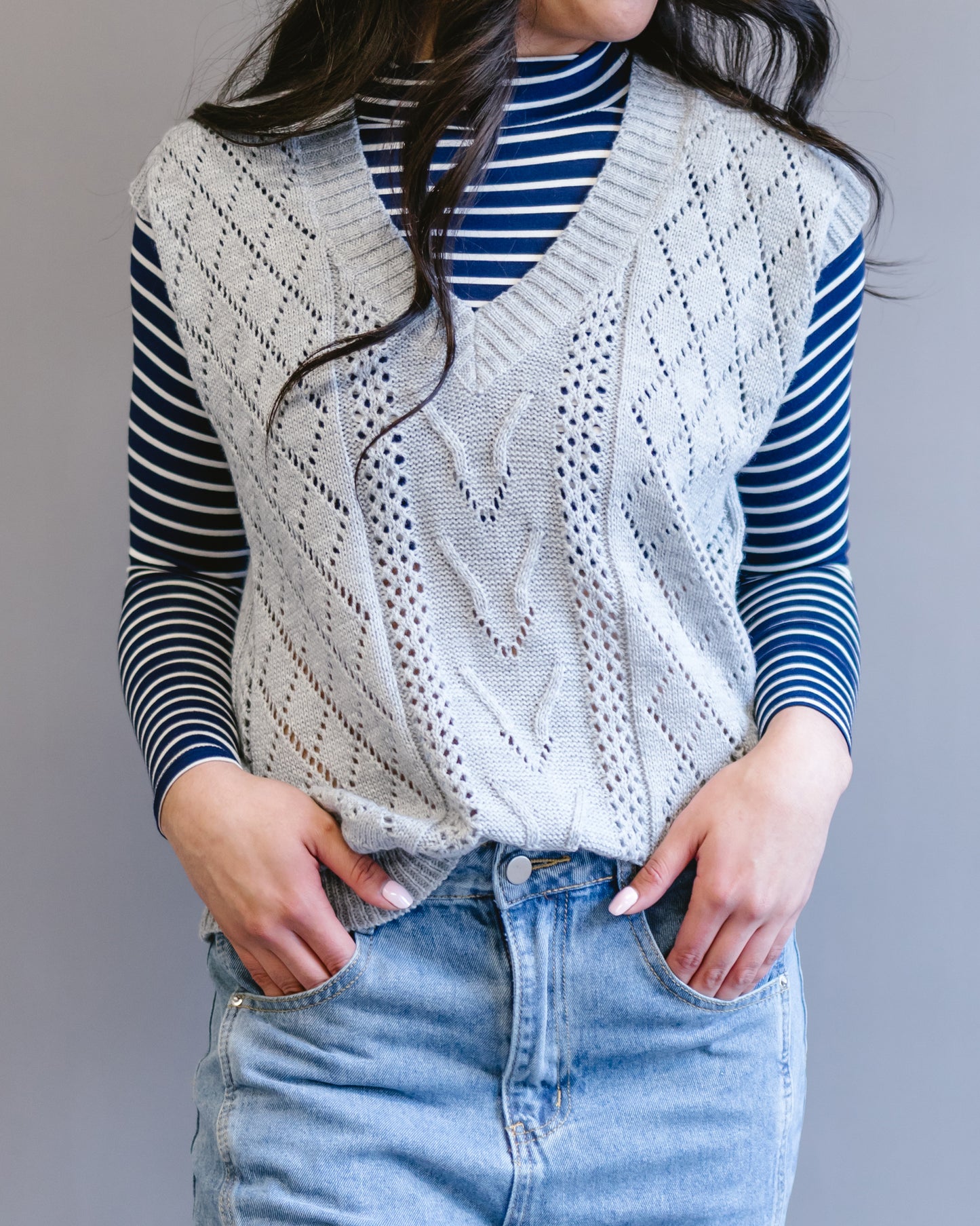 Woman wearing a gray open-knit sweater vest over a blue and white striped turtleneck and light blue jeans.