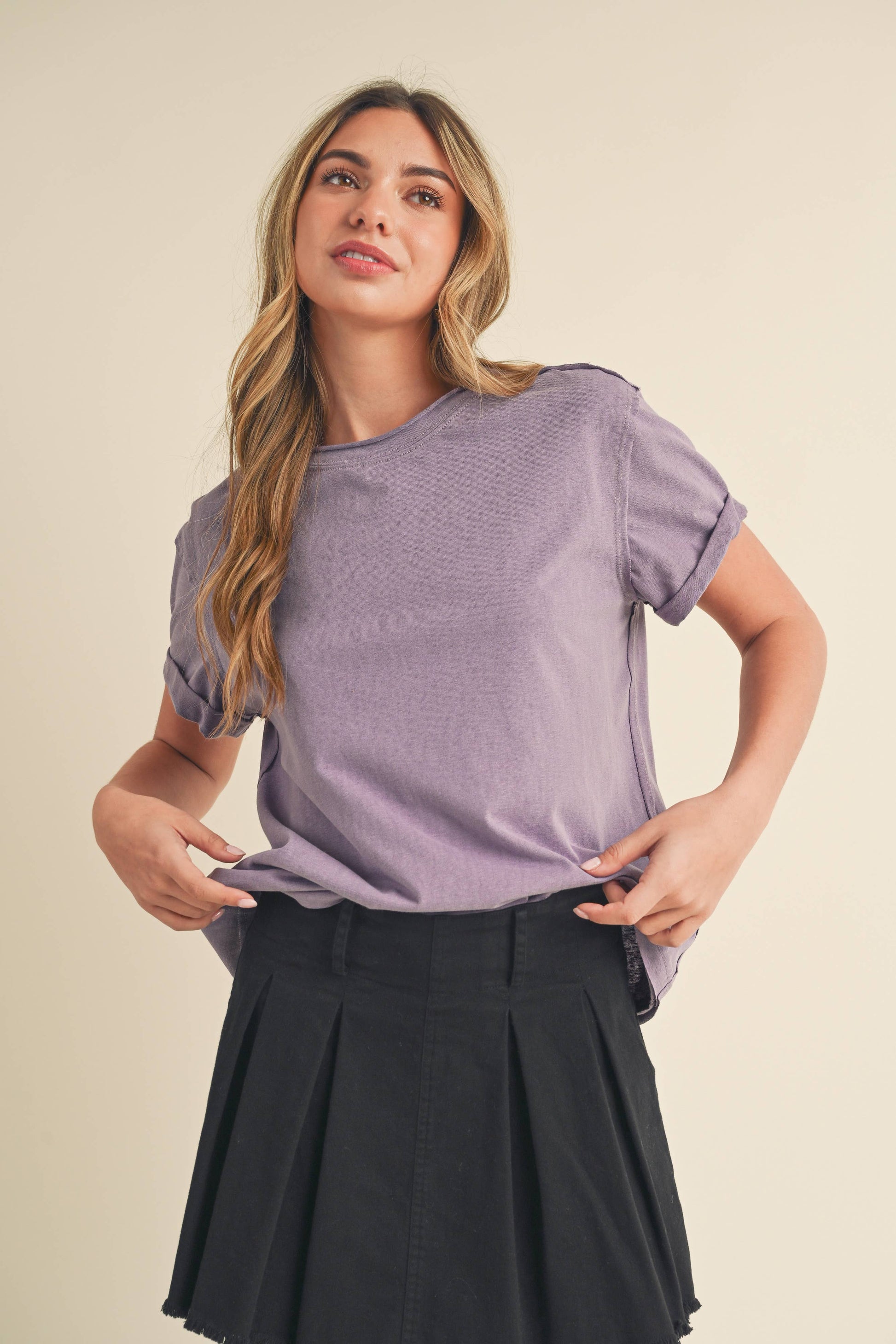 Young woman with long wavy hair, wearing a lavender t-shirt and a black pleated skirt, stands against a neutral background, exuding a relaxed vibe.
