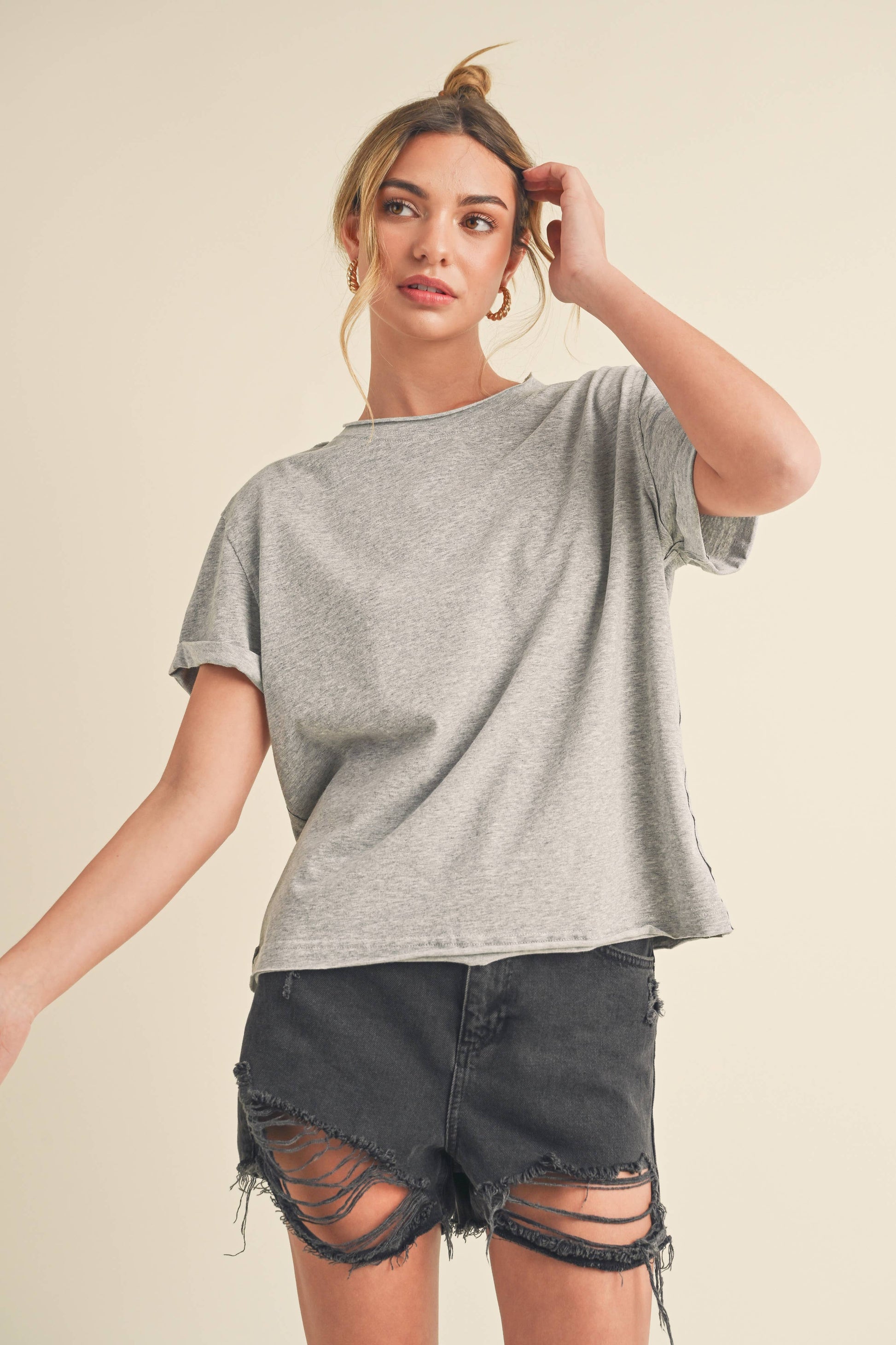 Young woman with a relaxed expression, wearing a loose gray T-shirt and distressed black denim shorts, stands against a light beige background.