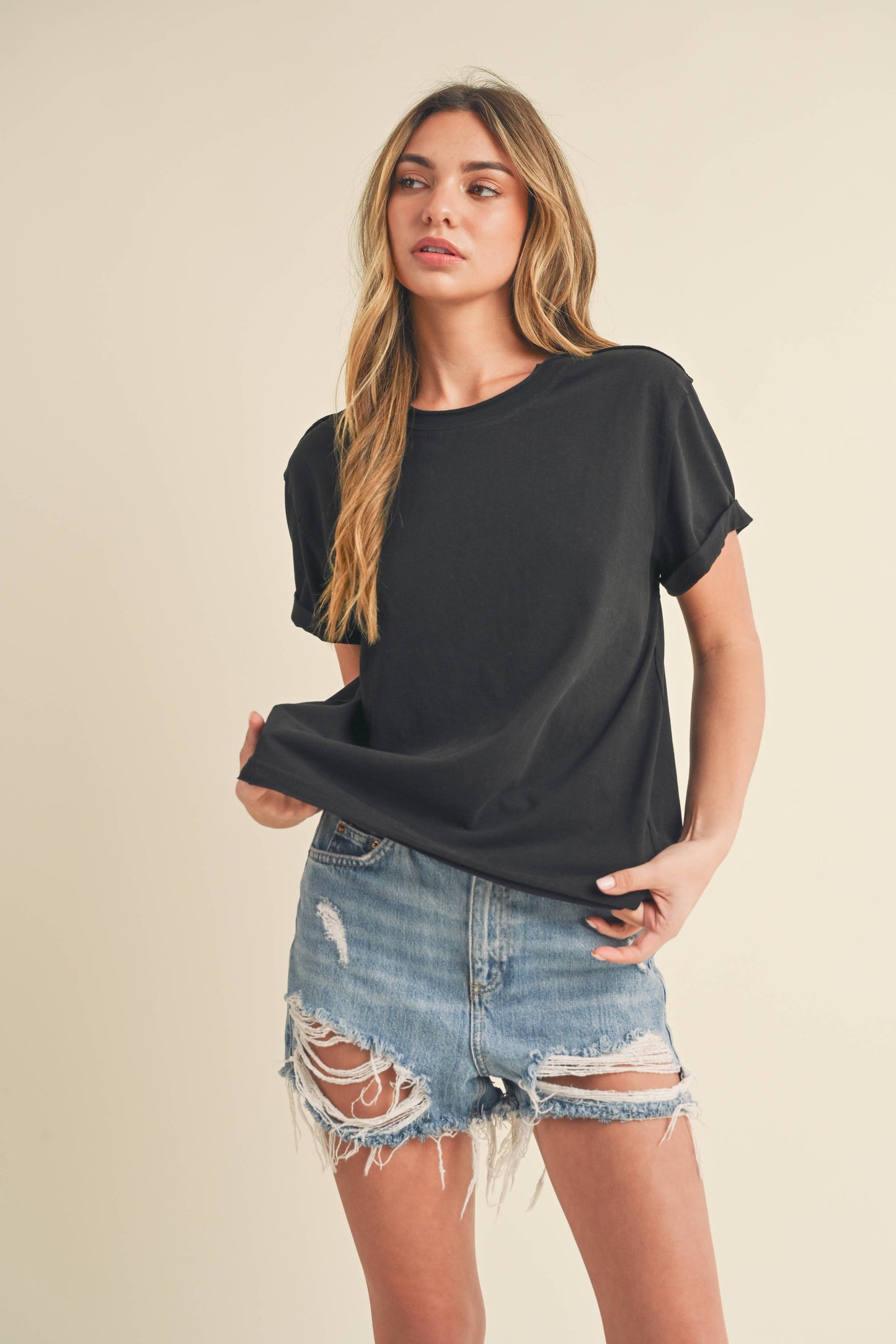 Young woman in a black T-shirt and distressed denim shorts stands against a beige background. She gazes slightly to the side with a neutral expression.