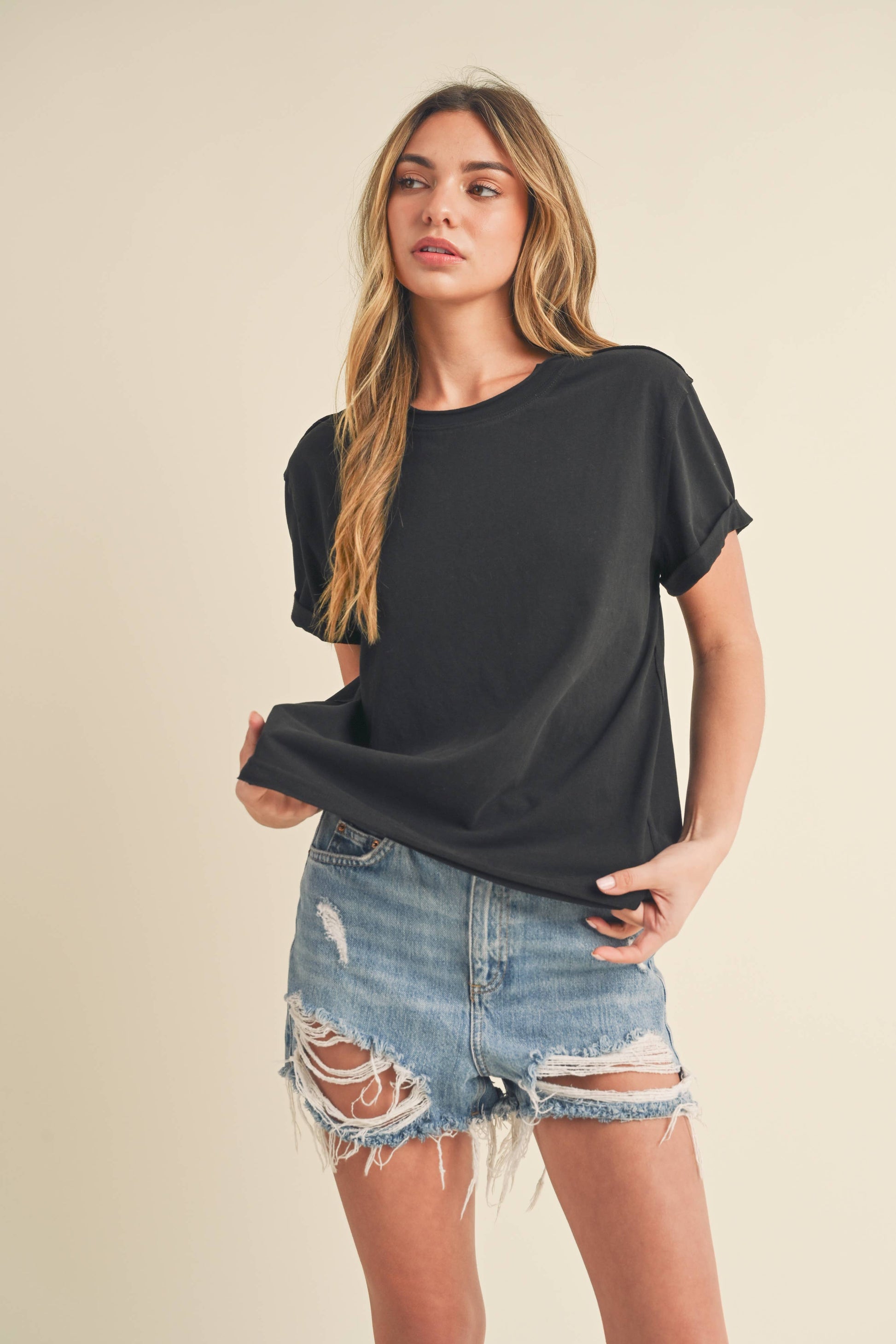 Young woman in a black T-shirt and distressed denim shorts stands against a beige background. She gazes slightly to the side with a neutral expression.