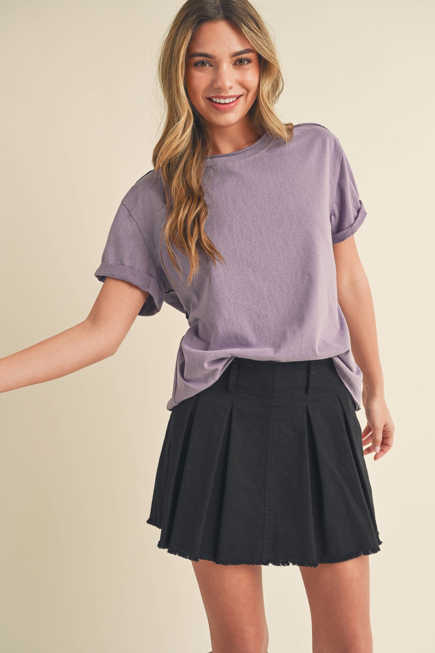 Smiling woman in a lavender t-shirt and black pleated skirt, standing against a beige background. Her long hair is loose, conveying a cheerful tone.