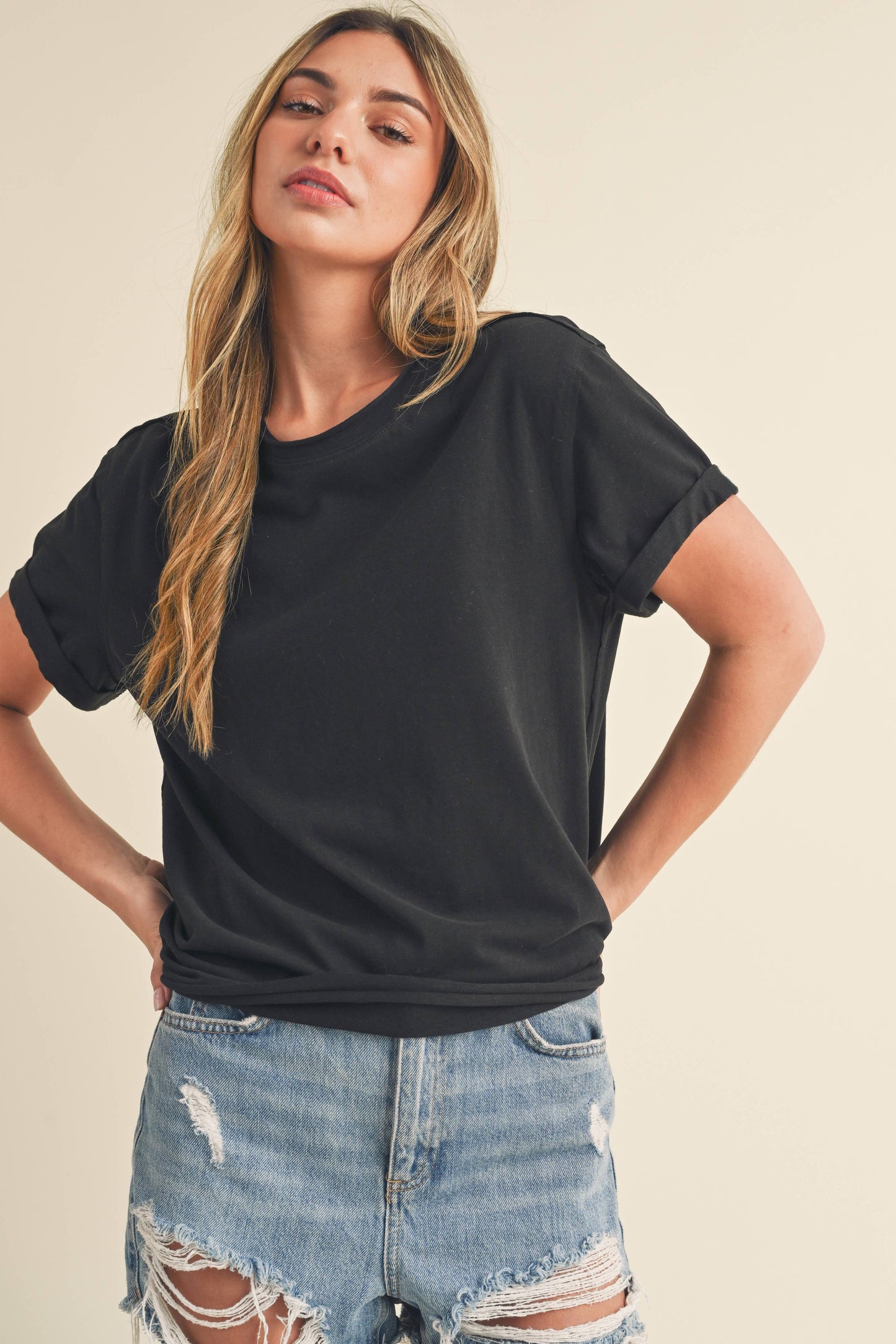 Young woman in a black t-shirt and distressed denim shorts stands confidently. Her long hair is loose, and she has a relaxed and laid-back expression.