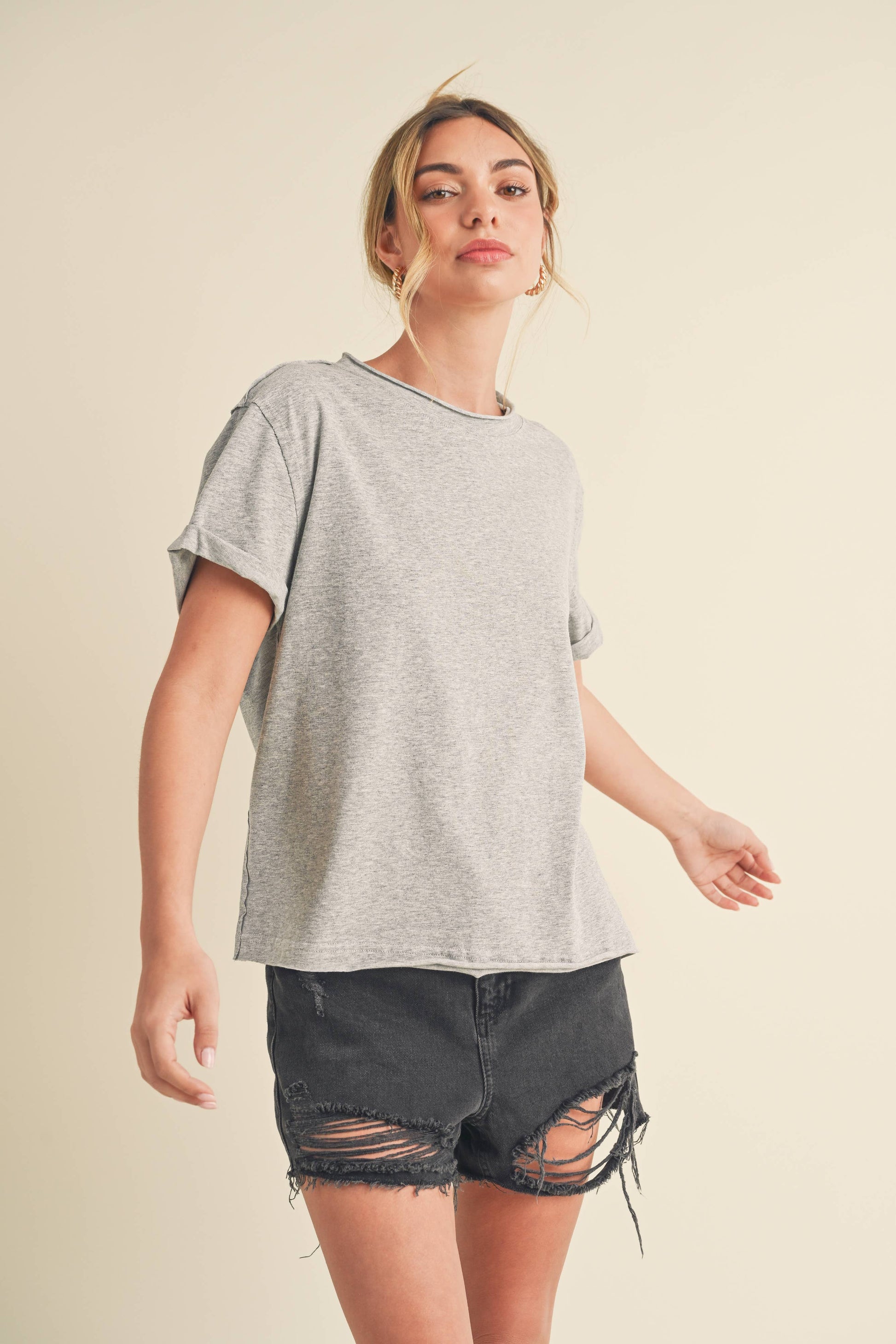 Woman with a relaxed pose wears a gray t-shirt and black distressed shorts, exuding a casual, laid-back vibe against a neutral backdrop.