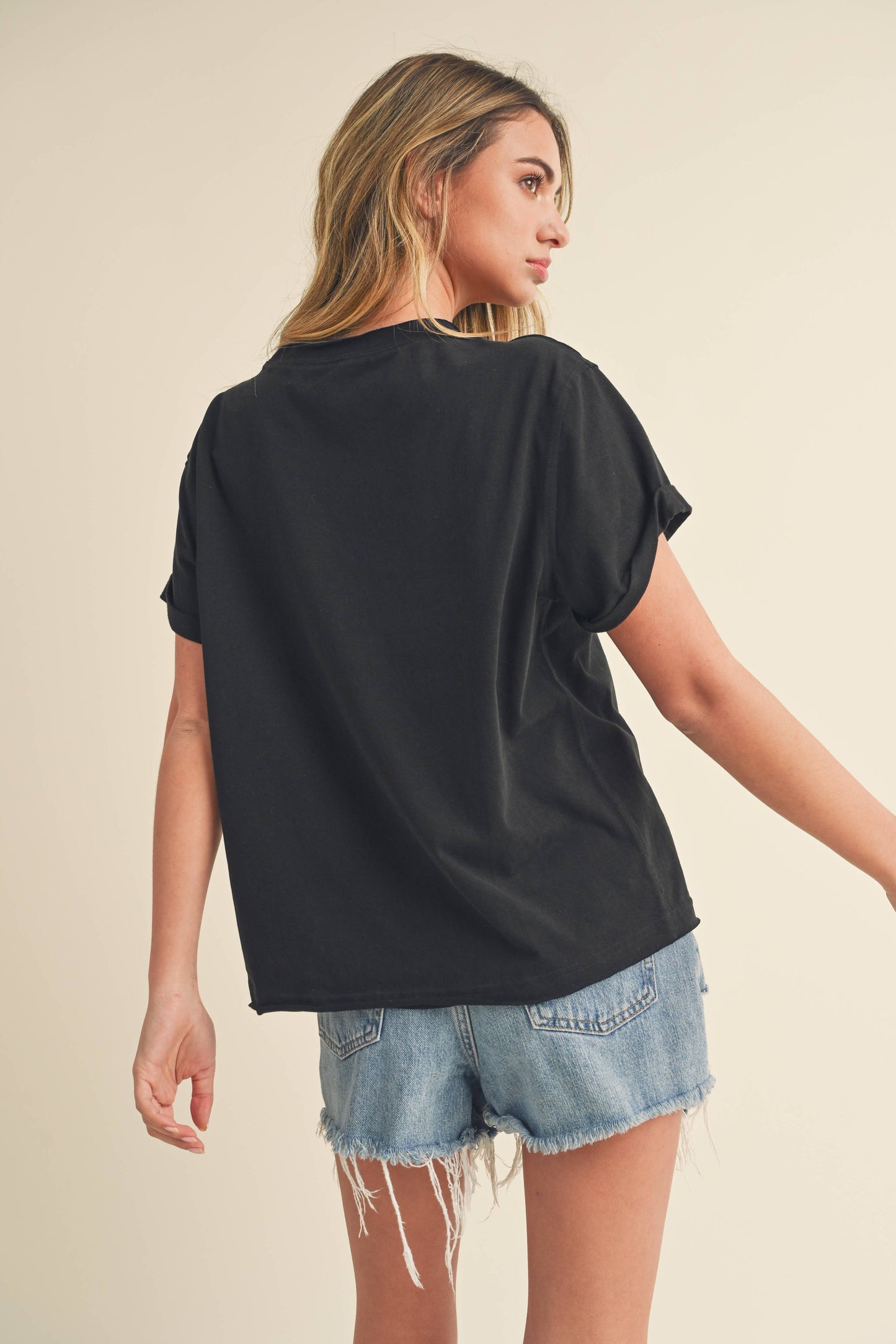 Young woman in a black T-shirt and frayed denim shorts, turned slightly away, exuding a casual, relaxed vibe against a neutral background.