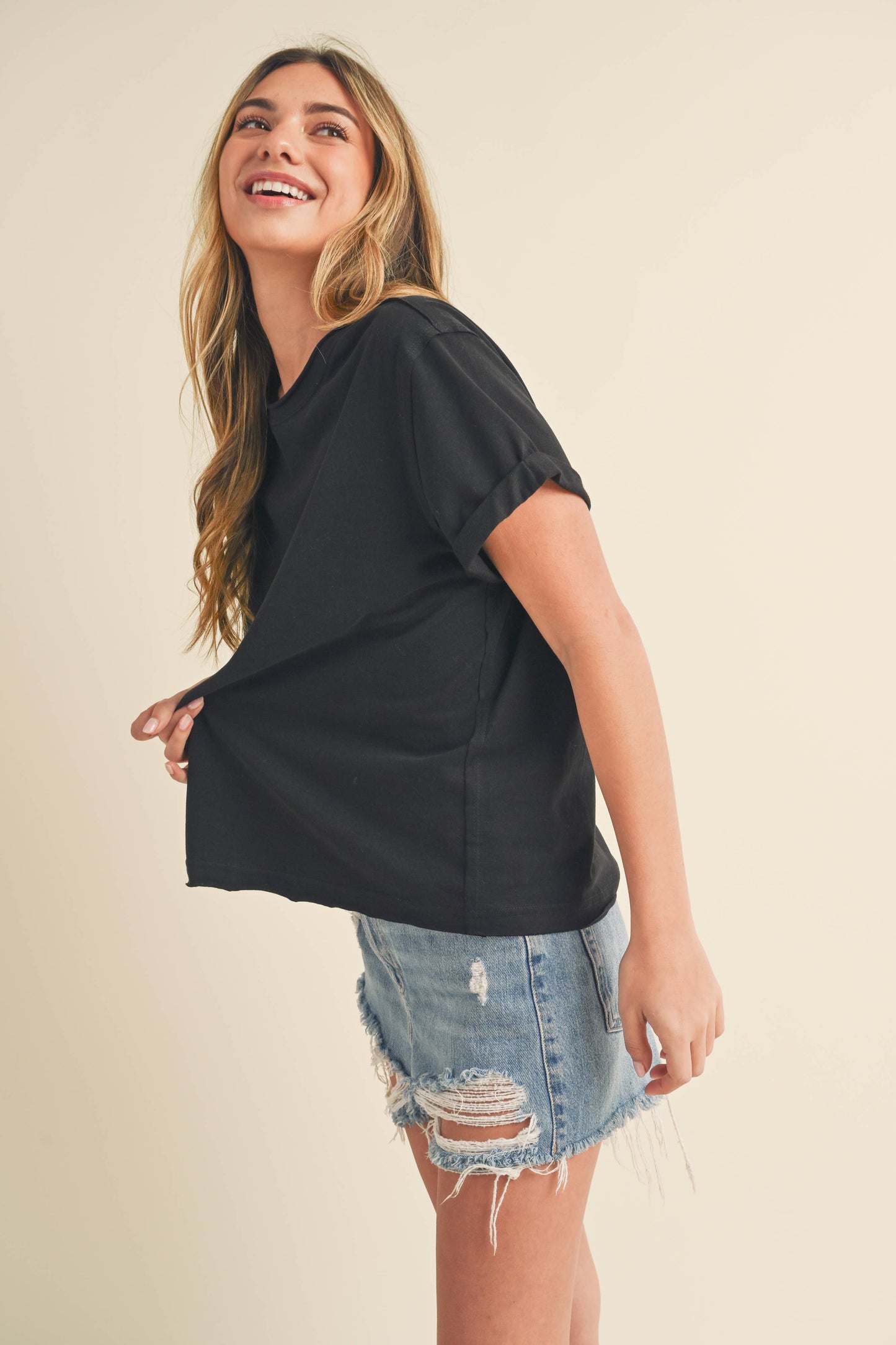 Smiling woman with long hair wearing a black t-shirt and distressed denim shorts, looking upwards. The background is a soft beige, conveying a casual, joyful tone.
