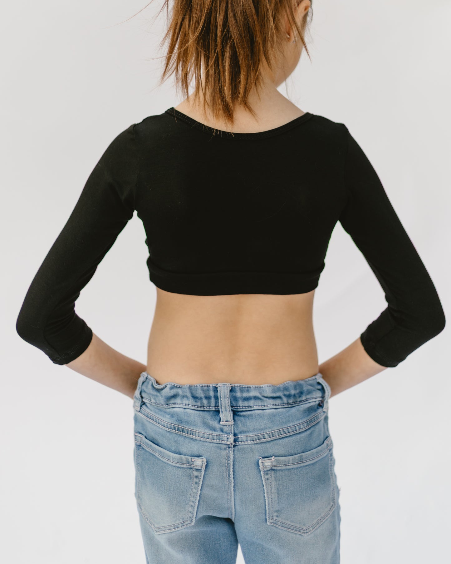 Young girl with ponytail, viewed from the back, wearing a black cropped top and high-waisted light blue jeans. Relaxed and casual setting.