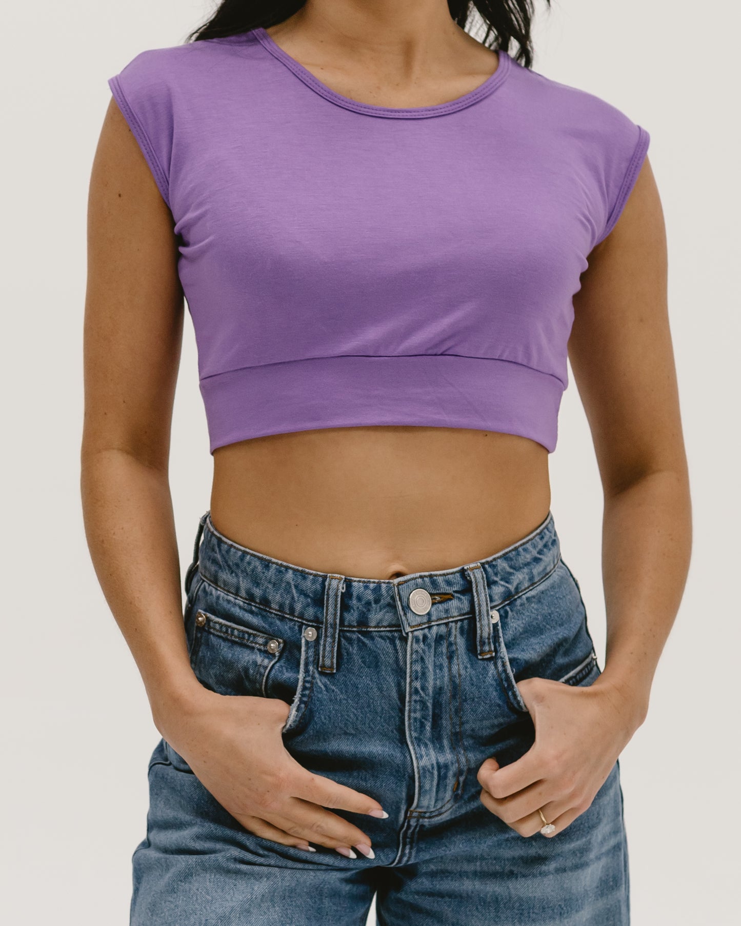 Back view of a woman wearing a purple Halftee crop top with short sleeves and blue denim jeans, showing her midriff and one hand on her hip