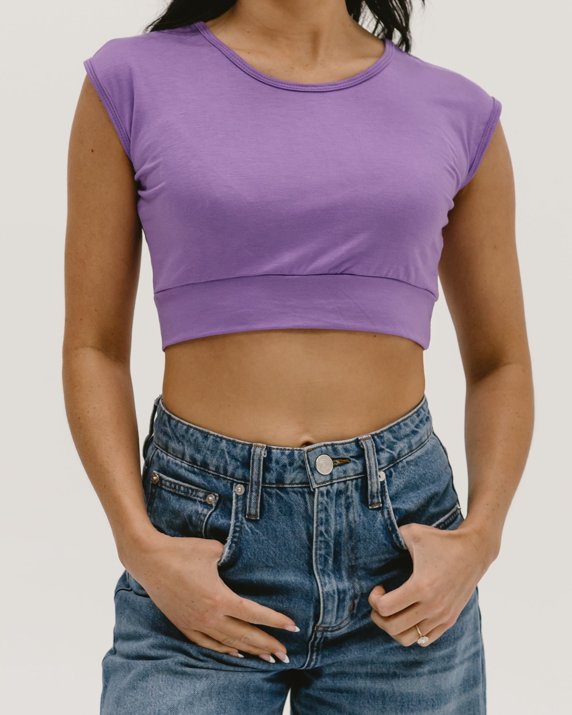 Back view of a woman wearing a purple Halftee crop top with short sleeves and blue denim jeans, showing her midriff and one hand on her hip