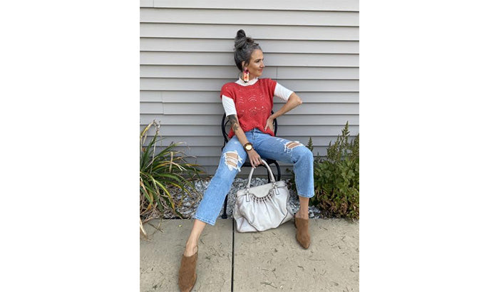 A person sitting on a chair against a gray wall, wearing a red sleeveless sweater over a white tee, distressed jeans, brown boots, and holding a white handbag, with plants in the background.