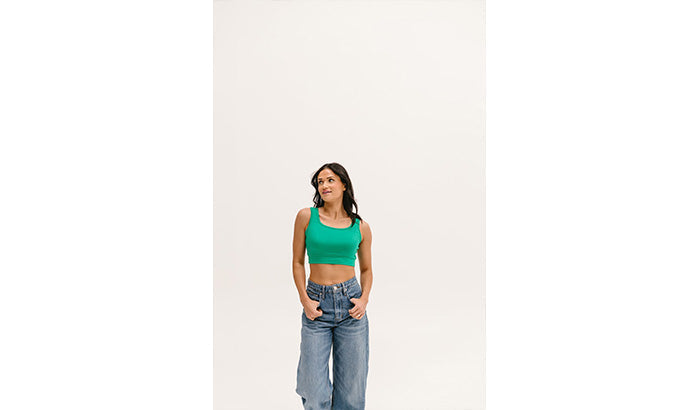 A woman in a teal crop top and jeans stands with hands in pockets against a white background.
