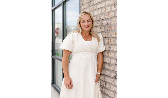 A woman in a white dress stands against a brick wall near a window.