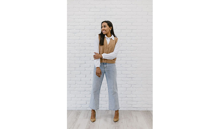 A woman in jeans and a vest stands against a white brick wall, showcasing her winter blouse and layered crew neck long sleeve.