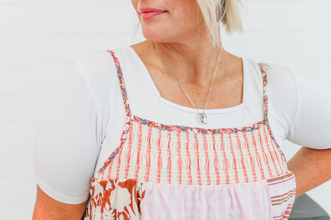 A woman wears a white tee under a patchwork pinafore dress with pink stripes and floral patterns, accessorized with a silver necklace.