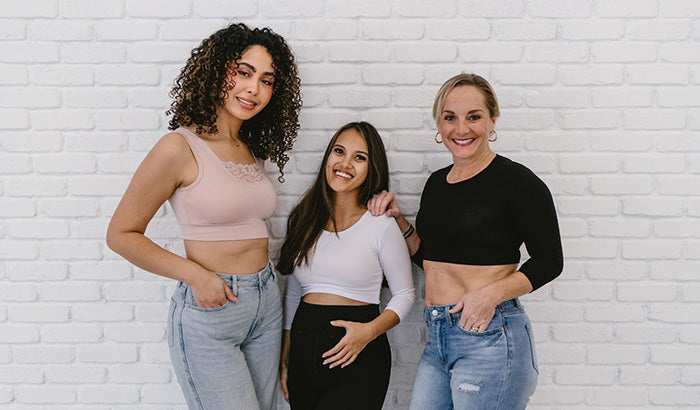 Three women in jeans and crop tops pose beside a brick wall, showcasing modesty shirts and festive Christmas deals.