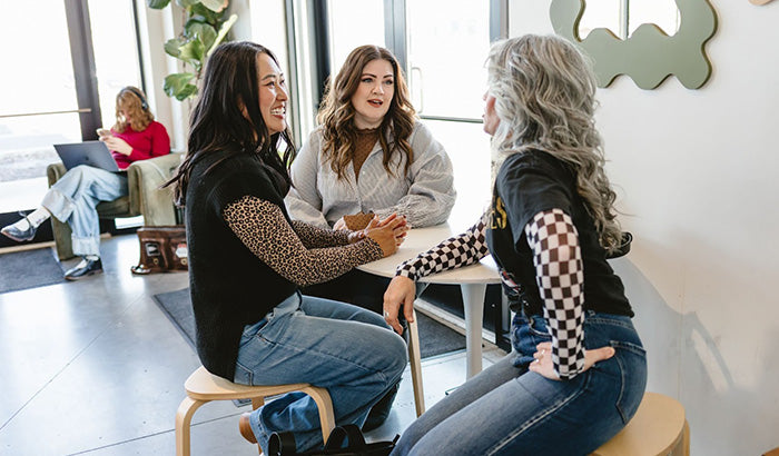 Three women in modest mid-sleeve shirts seated at a table in an office setting, engaged in discussion.