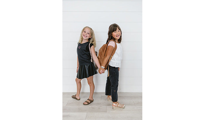 Two little girls in everyday skirts with mesh sleeves hold hands, standing together in front of a white wall.