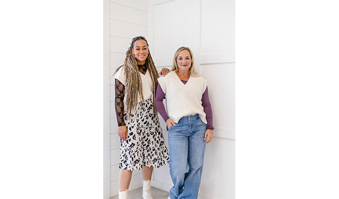 Two women in stylish sweater vests stand confidently in front of a white wall, showcasing a layered outfit trend.