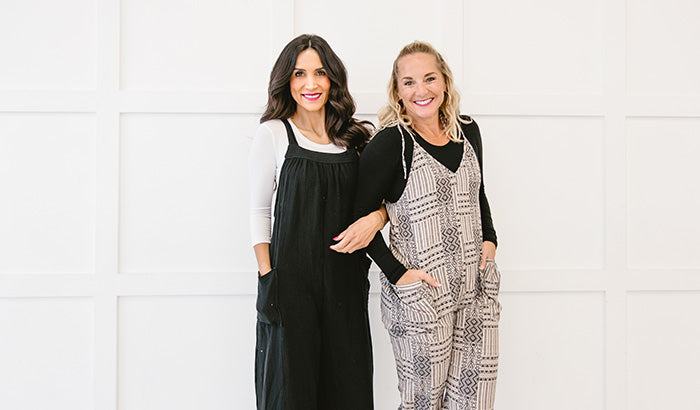Two Women In Overalls, Standing Against A White Wall. They Are Wearing Layered Shirts And Undershirts