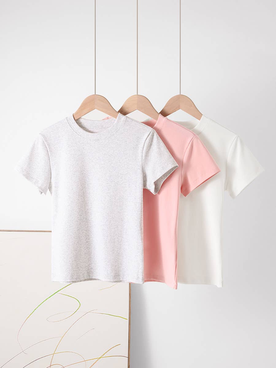 Three T-shirts in gray, pink, and white hang on wooden hangers against a minimal white background. A simple abstract painting is partially visible.