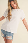 A woman in a casual pose wears a white T-shirt and distressed denim shorts. Her long, wavy hair frames her face as she looks downward, creating a relaxed and effortless vibe against a neutral background.