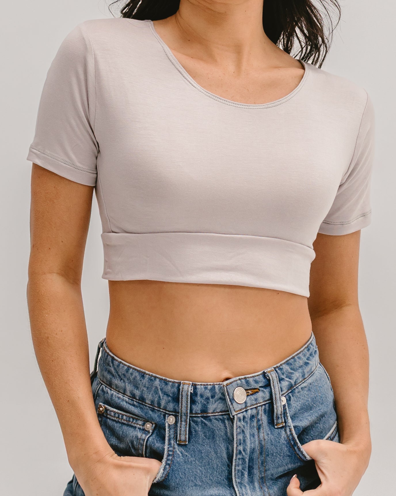 A cropped view of a person wearing a white three-quarter sleeve Halftee and high-waisted light-wash jeans, holding the hem of the Halftee with both hands.