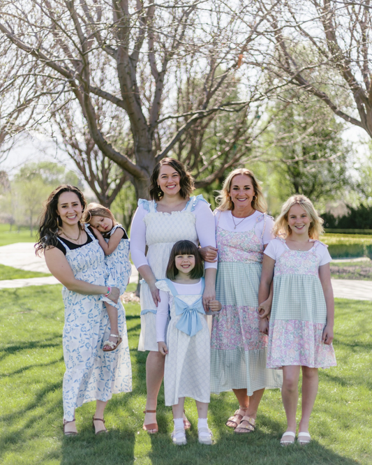 A group of five women and girls stand on a grassy lawn under leafless trees. They are smiling, wearing patterned sundresses, exuding a warm and joyful atmosphere.