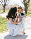 A woman and child in matching blue floral dresses are outside on a sunny day, sharing a moment. The setting is a park with green trees and grass.