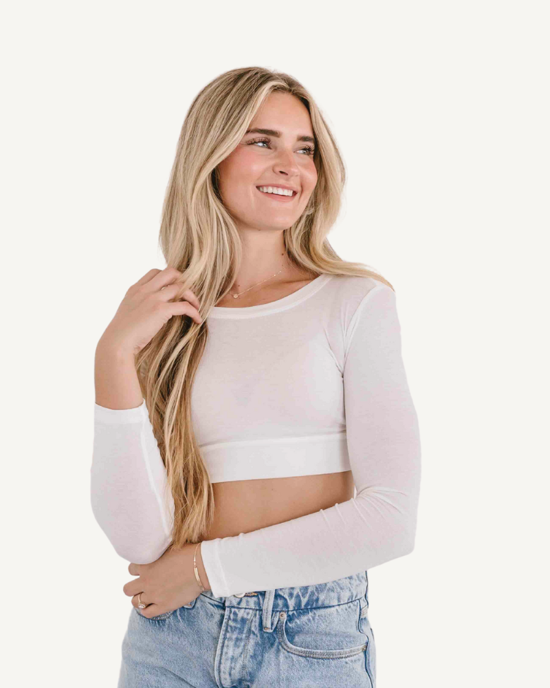 A woman wearing a white crop top and jeans, with a crew neck long sleeve.