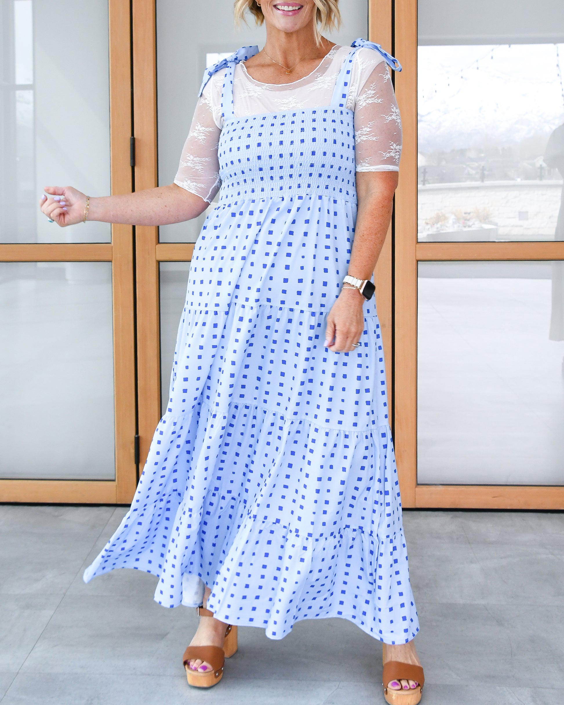 Woman smiling and twirling in a blue and white checkered sundress over a white sheer top and brown platform sandals.