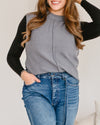 Happy person in a charcoal gray knit vest layered over a black long-sleeved shirt and light wash denim jeans.