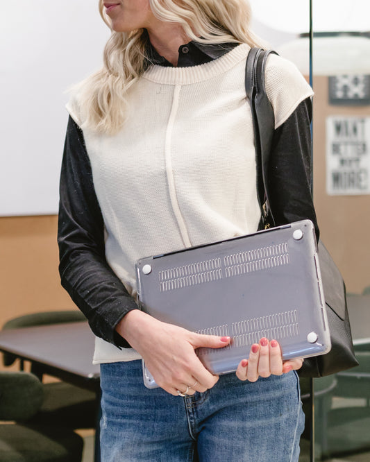 Torso shot of a woman holding a closed laptop, wearing a cream sweater vest with black sleeves and collar, blue jeans, and carrying a black shoulder bag.