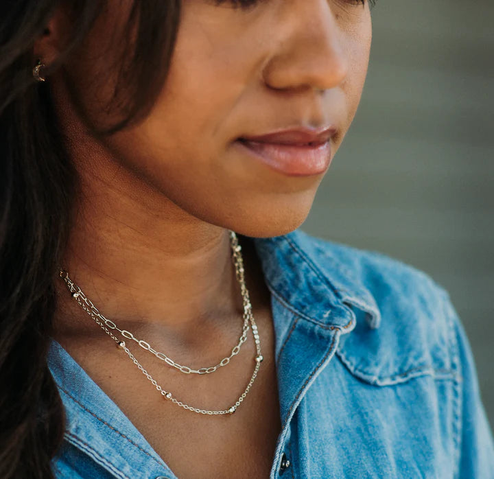 A woman in a green and red sweater sitting on a couch, accessorized with necklaces.