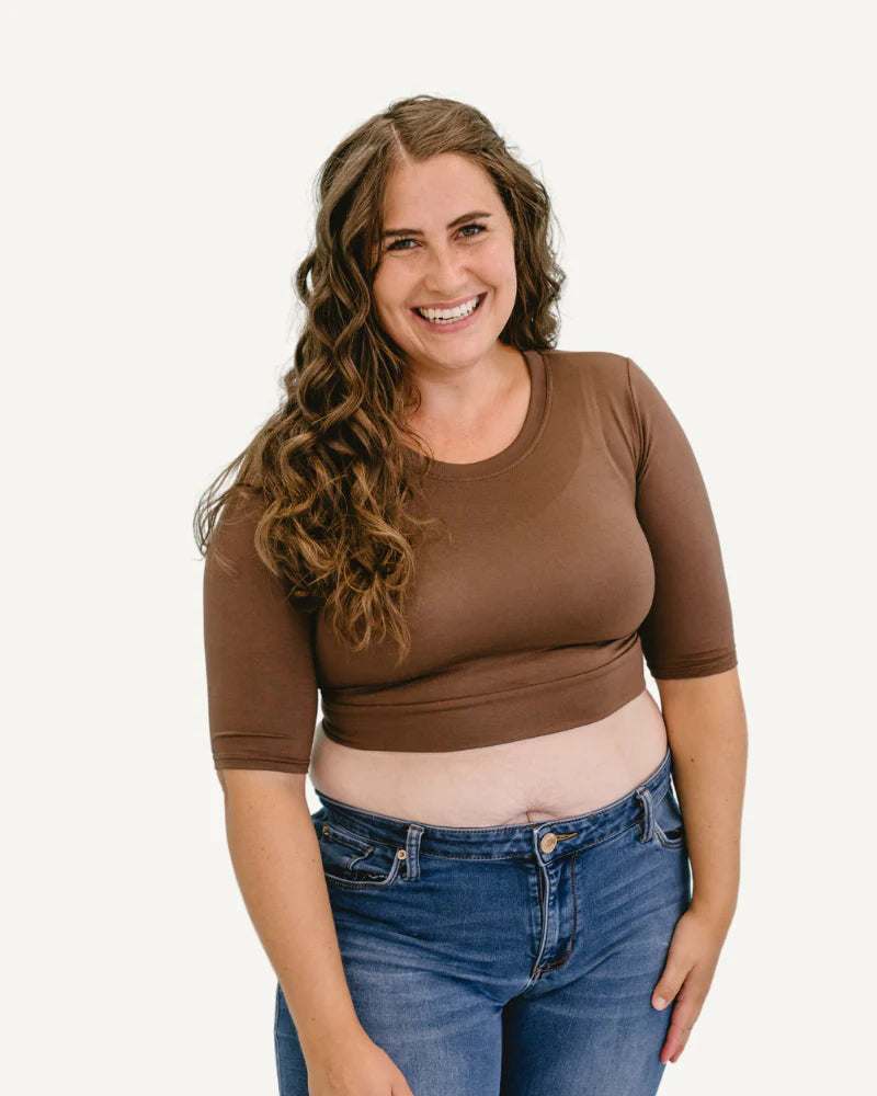 A woman wearing a brown top and jeans stands confidently, showcasing a casual yet stylish outfit.