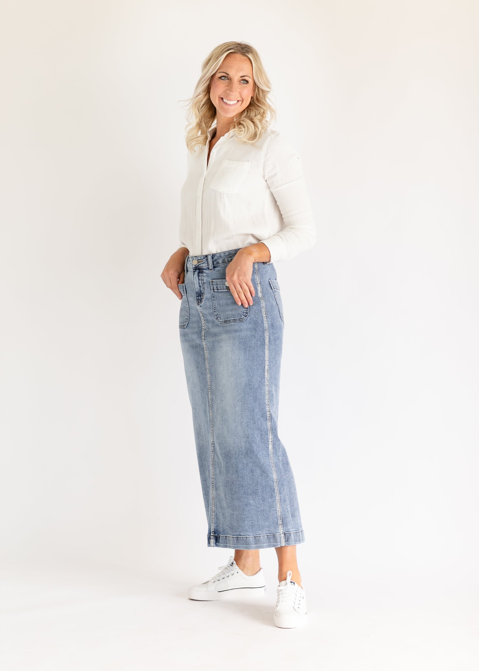 Smiling woman in a white blouse and denim skirt stands against a plain white background, exuding a casual and cheerful vibe.