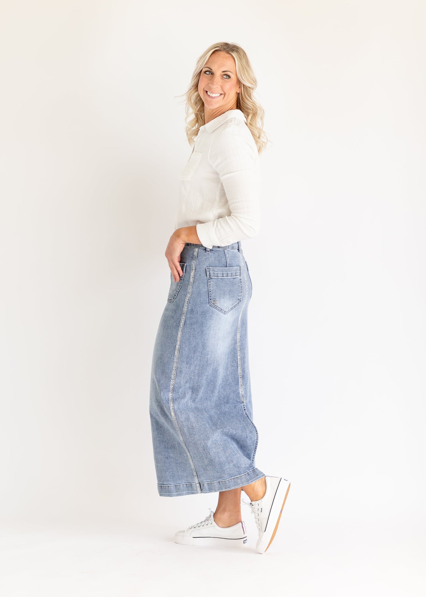 A smiling person stands sideways in a white shirt and long denim skirt, wearing white sneakers. The background is plain white, creating a bright, casual feel.